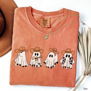 May include: A burnt orange t-shirt with a graphic of four ghosts wearing cowboy hats. The ghosts are playing musical instruments. The text "COMFORT COLORS" is on the tag.