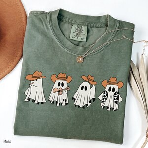 May include: A green t-shirt with a graphic of four ghosts wearing cowboy hats. One ghost is playing a ukulele. The text "COMFORT COLORS" is visible on the tag.
