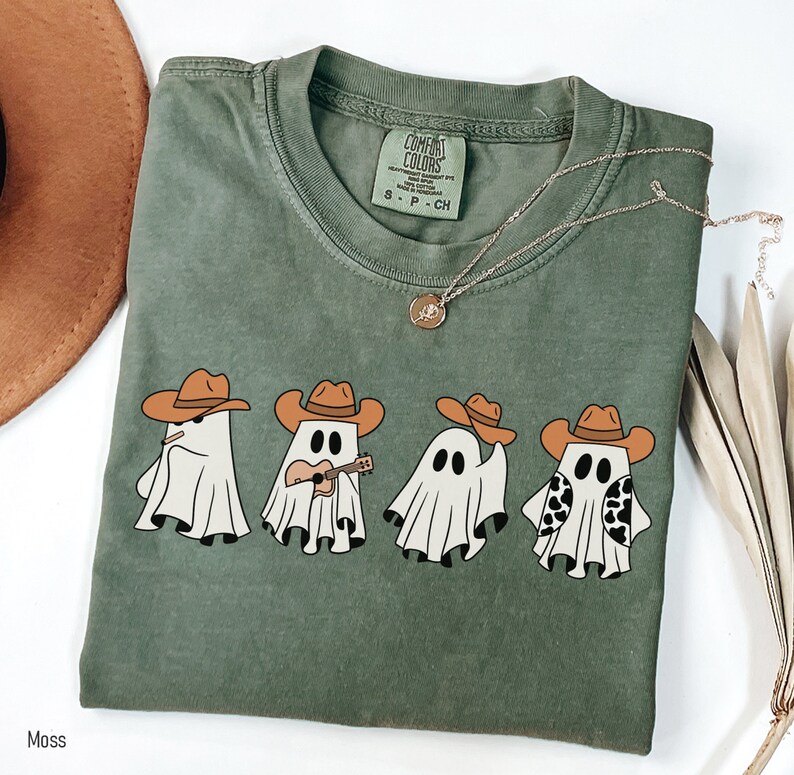 May include: A green t-shirt with a graphic of four ghosts wearing cowboy hats. One ghost is playing a ukulele. The text "COMFORT COLORS" is visible on the tag.