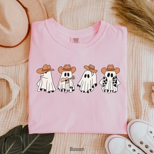 May include: Pink t-shirt with a graphic of four ghosts wearing cowboy hats. One ghost is playing a guitar. The text "COMFORT COLORS" is visible on the tag.