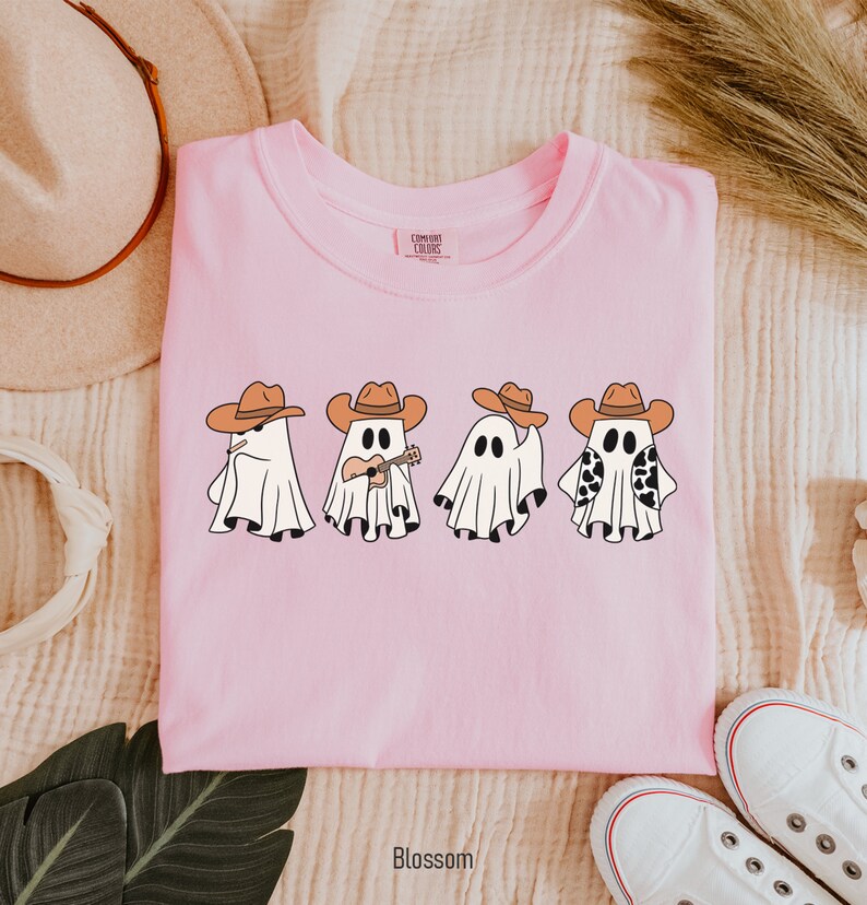 May include: Pink t-shirt with a graphic of four ghosts wearing cowboy hats. One ghost is playing a guitar. The text "COMFORT COLORS" is visible on the tag.