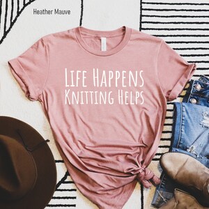 May include: A heather mauve t-shirt with the text "Life Happens Knitting Helps" printed in white.