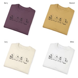 May include: Four white ghost illustrations with stacks of books are printed on a t-shirt. The t-shirt is available in four colors: berry, mustard, ivory, and white.