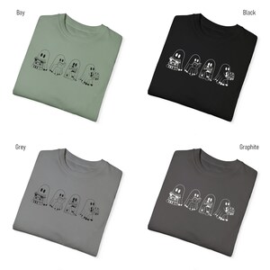 May include: Four white outline ghosts wearing hats and holding stacks of books on a t-shirt. The t-shirt is available in four colors: bay, black, grey, and graphite.