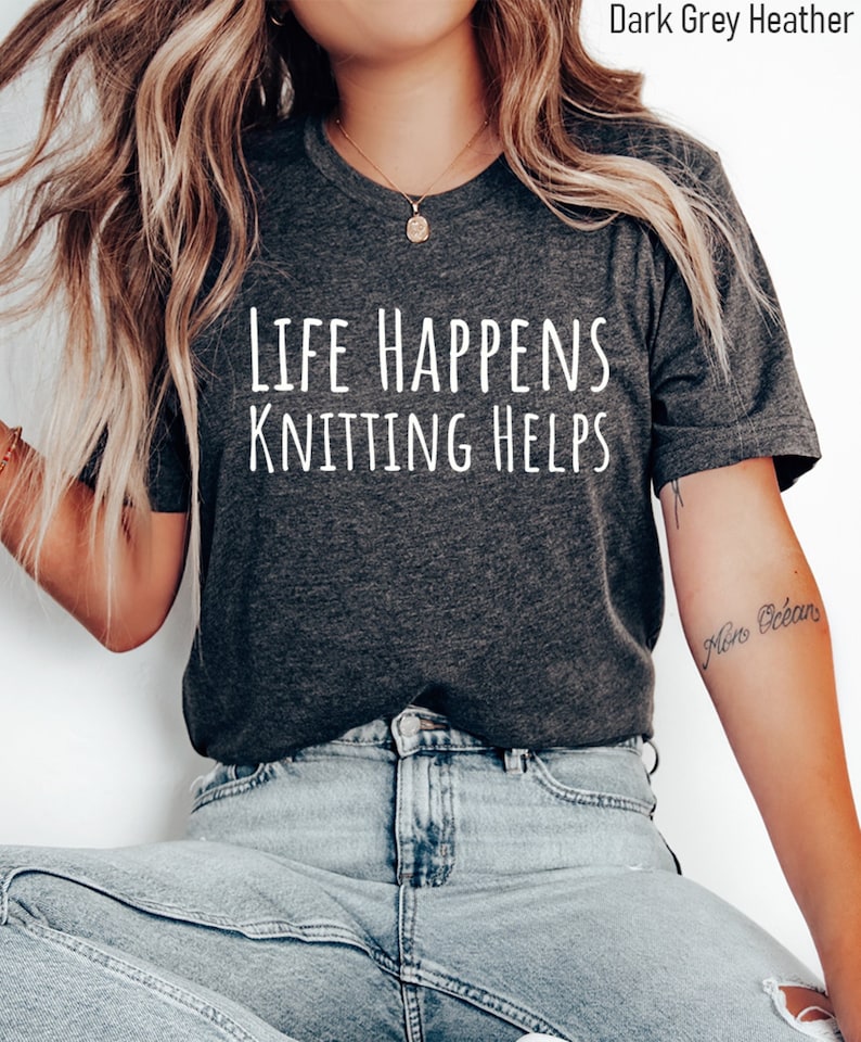 May include: A dark grey heather t-shirt with the text "Life Happens Knitting Helps" printed in white.