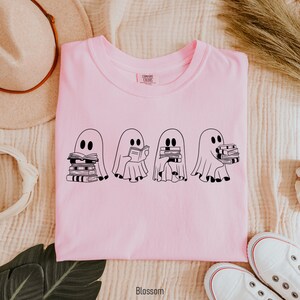 May include: Pink t-shirt with a graphic of four ghosts reading books. The ghosts are black and white and are holding stacks of books. The text "COMFORT COLORS" is visible on the tag.