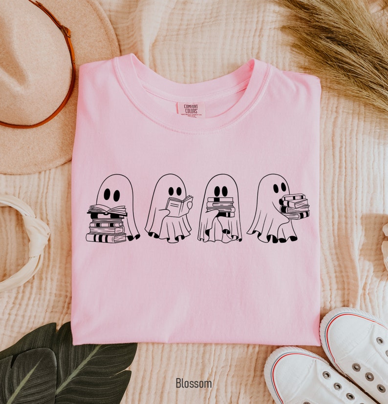 May include: Pink t-shirt with a graphic of four ghosts reading books. The ghosts are black and white and are holding stacks of books. The text "COMFORT COLORS" is visible on the tag.