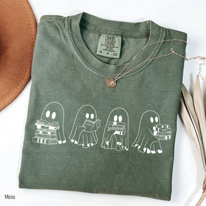May include: A green t-shirt with a white graphic of four ghosts reading books. The text "COMFORT COLORS" is on the tag. The shirt is folded and laid on a white surface.