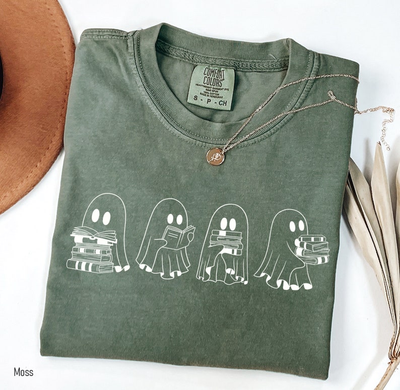 May include: A green t-shirt with a white graphic of four ghosts reading books. The text "COMFORT COLORS" is on the tag. The shirt is folded and laid on a white surface.