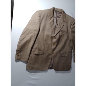 May include: A tan blazer with a subtle plaid pattern. The jacket features a notched lapel, three-button closure, and two front pockets. The sleeves have four buttons at the cuff. The blazer is made of a textured fabric.