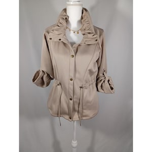 May include: A tan jacket with a high, ruffled collar and three-quarter sleeves. The jacket features a drawstring waist, gold-colored buttons, and rolled-up sleeves. The jacket is displayed on a white mannequin.