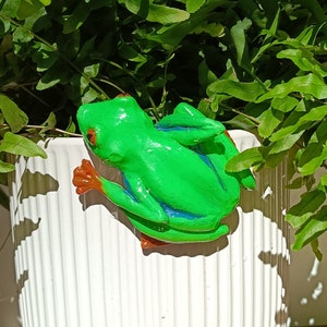 Puede incluir: Una vibrante figura de rana verde con ojos rojos y patas naranjas, posada sobre una maceta blanca con estrías verticales. La rana tiene detalles azules y está rodeada de exuberante follaje verde.