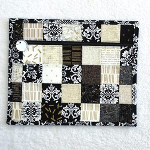 Peut inclure: Une pochette matelassée rectangulaire avec une fermeture éclair noire et une étiquette circulaire blanche. Le motif patchwork présente des carrés noirs, blancs et dorés avec des motifs floraux et géométriques. La pochette mesure environ 25 cm de large et 18 cm de haut.