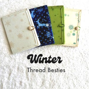 May include: Four rectangular fabric needle cases with button and elastic closures. The designs include cream with gold stars, dark blue with white deer and snowflakes, solid green with starbursts, and cream with green snowflakes. Text reads Winter Thread Besties.