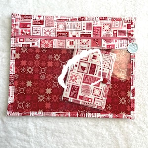 Può includere: Una pochette natalizia rossa e bianca con un pannello frontale trasparente. Presenta un motivo festivo di francobolli, fiocchi di neve e testo come "Holly Jolly" e "A Quilty Little Christmas". È attaccato un tag più piccolo con un design abbinato.