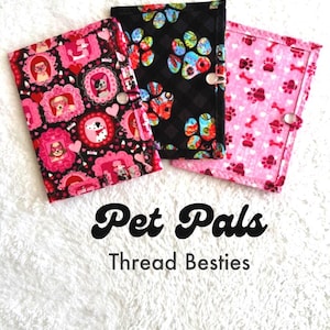 May include: Three rectangular fabric cases with snap closures. The left case features pink frames with dogs on a black background. The center case has colorful paw prints on a black background. The right case displays pink paw prints and bones.