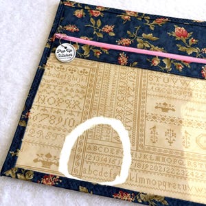 Può includere: Una borsa per progetti di punto croce con un bordo a stampa floreale blu navy e un pannello frontale trasparente. La borsa ha una cerniera rosa e un ciondolo in metallo con la scritta "Pop Up Stitcher". Il tessuto interno presenta un motivo alfabetico vintage.