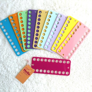 Puede incluir: Una colección de organizadores de hilo de plástico de colores en azul, naranja, verde, morado, amarillo y rosa. Cada organizador tiene una serie de agujeros y un recorte en forma de corazón. También hay un organizador magenta y una etiqueta que dice "Hello handmade".