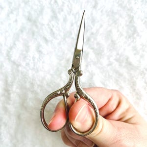 May include: Silver-toned embroidery scissors with ornate handles and sharp, pointed blades. The scissors have a classic design, ideal for detailed needlework and crafting projects. The blades are approximately 1.5 inches long.