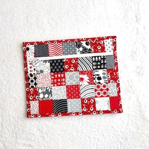 May include: A rectangular patchwork pouch with a red border and a white zip. The patchwork design features squares in red, white, grey, and black with floral, striped, and geometric patterns. The pouch is on a white surface.