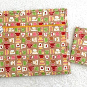 Può includere: Un astuccio in tessuto beige con cerniera e una piccola custodia per taccuino abbinata. Entrambi gli articoli presentano un motivo ripetuto di tazze colorate in rosso, verde, rosa e giallo. L'astuccio ha una cerniera gialla e un piccolo bottone rotondo.