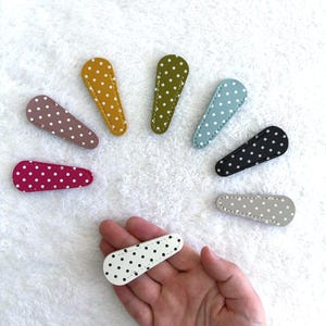 Puede incluir: Colección de pinzas para el pelo con lunares en varios colores, como amarillo mostaza, verde oliva y burdeos. Una pinza se sostiene en una mano, mostrando la base blanca y los lunares negros. Las pinzas están dispuestas en un patrón circular.