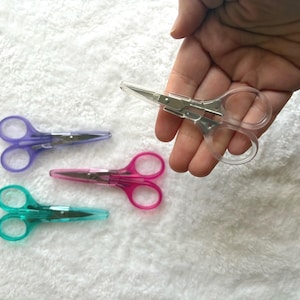 May include: A set of small, colorful scissors with clear plastic handles and stainless steel blades. The scissors are in shades of purple, pink, and teal. One pair is held in a hand, while the others are arranged on a white, textured surface.