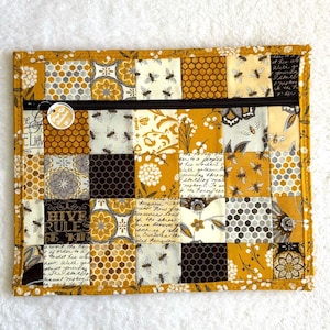 Può includere: Pochette trapuntata con un design patchwork con motivi a tema api e favi in tonalità giallo, marrone e crema. La pochette ha una cerniera nera e un'etichetta rotonda. Il testo "HIVE RULES" è visibile.