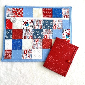 Può includere: Una custodia rettangolare con cerniera caratterizzata da un design patchwork di quadrati di tessuto rossi, bianchi e blu con testi e motivi patriottici, abbinata a un piccolo taccuino rivestito in tessuto rosso con chiusura a bottone e stelle.