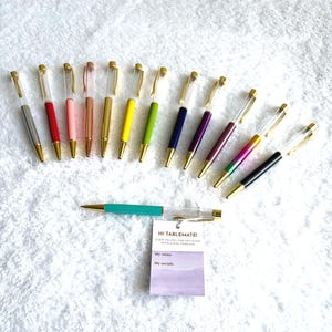 May include: A collection of colorful pens with gold-colored accents and clear barrels. The pens are arranged on a white, textured surface. One pen has a teal barrel. A small tag reads "HI TABLEMATE!" with space for a name and social media.