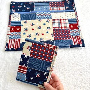 Può includere: Una custodia in tessuto rettangolare con chiusura a zip e una copertina per quaderno in tessuto abbinata, entrambe caratterizzate da un design patchwork con stelle, strisce e motivi geometrici rossi, bianchi e blu. La custodia ha un'etichetta.