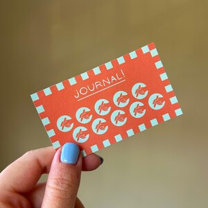 以下が含まれることがあります： オレンジ色の長方形のカードで、白で「JOURNAL！」と書かれています。カードには、水色の手のイラストが繰り返されています。ボーダーは水色の正方形です。