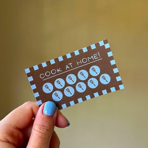 以下が含まれることがあります： 「COOK AT HOME!」の文字と、水色の丸の中にヘラのアイコンが描かれた茶色のカード。カードの縁は茶色と水色の正方形が交互に並んでいます。水色のマニキュアを塗った手でカードを持っています。