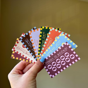 以下が含まれることがあります： さまざまなフレーズと本のアイコンが描かれた、カラフルなカードの扇子を手に持っています。カードには市松模様のボーダーがあり、「SOMETHING!」、「AT HOME!」、「ME WITH KIDS!」、「SPEND DAY!」、「JOURNAL!」、「SPREAD JOY!」、および「FINISH 10 BOOKS!」と書かれています。