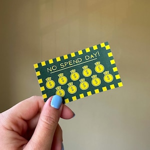 以下が含まれることがあります： ドル記号の入ったお金の袋が描かれた、緑と黄色の「No Spend Day」スクラッチカード。カードには黄色のチェック柄の縁取りがあり、青いマニキュアを塗った人が持っています。