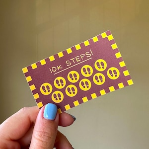 以下が含まれることがあります： 濃い赤色の背景と黄色の縁取りの長方形のカード。カードには「10K STEPS！」の文字と黄色の足跡が描かれています。青いマニキュアを塗った手でカードを持っています。