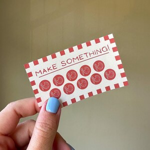 以下が含まれることがあります： 赤と白のチェック柄の縁取りと「MAKE SOMETHING！」の文字が黒で印刷された長方形のカード。カードには、筆とパレットのデザインが施された赤い円が描かれています。青いマニキュアを塗った手でカードが持たれています。