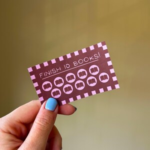 以下が含まれることがあります： 「FINISH 10 BOOKS！」の文字と10冊の本のアイコンが描かれたピンクと茶色のカード。カードにはチェック柄のボーダーがあり、青いマニキュアを塗った手で持たれています。