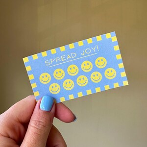 以下が含まれることがあります： 「SPREAD JOY！」の文字と黄色のスマイリーフェイスが描かれた青いカード。カードには黄色のチェック柄のボーダーがあります。青いマニキュアを塗った人がカードを持っています。