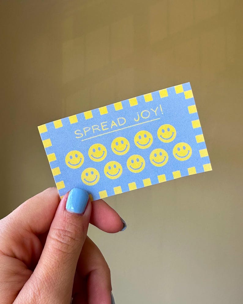 以下が含まれることがあります： 「SPREAD JOY！」の文字と黄色のスマイリーフェイスが描かれた青いカード。カードには黄色のチェック柄のボーダーがあります。青いマニキュアを塗った人がカードを持っています。