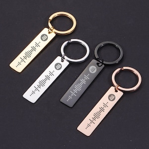 Könnte beinhalten: Vier Schlüsselanhänger mit verschiedenen Metalloberflächen: Gold, Silber, Schwarz und Roségold. Jeder Schlüsselanhänger hat ein rechteckiges Schild mit einem Spotify-Logo und einem Barcode-ähnlichen Design.