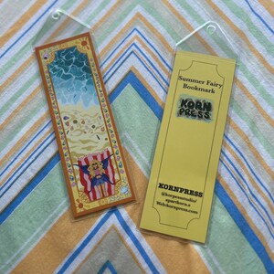 May include: Two bookmarks in clear plastic sleeves. One depicts a beach scene with a fairy, the other is yellow with the text "Summer Fairy Bookmark" and the "Korn Press" logo. The bookmarks are on a striped background.