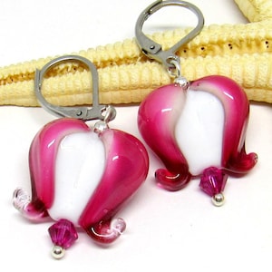 Könnte beinhalten: Ein Paar rosa und weiße Glasblumenohrringe mit silbernen Haken. Die Ohrringe sind glockenförmig mit einem weißen Zentrum und einer rosa Außenhülle. Jeder Ohrring hat einen kleinen rosa Kristall, der an der Unterseite hängt.