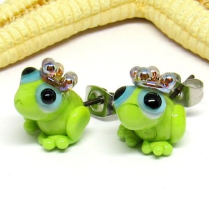 Könnte beinhalten: Ein Paar grüne Frosch-Ohrringe mit schillernden Kronen und blauen Augen. Die Ohrringe sind aus Glas und haben Edelstahl-Stifte.