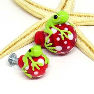 Peut inclure: Deux figurines de grenouilles en verre vert perchées sur des boutons en forme de champignon en verre rouge et blanc à pois. Les boutons sont utilisés pour les poignées de placard ou de tiroir.
