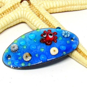 Könnte beinhalten: Eine blaue, ovale Emaille-Anstecknadel mit einem roten Oktopus mit weißen Augen und weißen und schwarzen Muscheln auf blauem Hintergrund mit grünen und blauen Punkten.