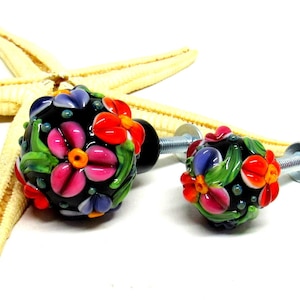 Könnte beinhalten: Zwei bunte Glas-Schrankknöpfe mit Blumendesigns. Die Knöpfe sind schwarz mit roten, rosa, orangefarbenen, gelben, grünen und blauen Blumen. Die Knöpfe sind an silbernen Metallschrauben befestigt.