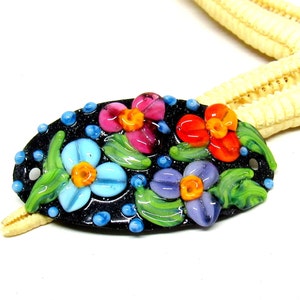 Könnte beinhalten: Eine schwarze ovale Glasperle mit einem Blumenmuster. Die Perle zeigt vier Blüten in den Farben Pink, Orange, Blau und Lila mit grünen Blättern und gelben Zentren. Die Perle ist mit kleinen blauen Punkten verziert.