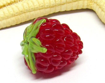 May include: A red glass raspberry bead with a green glass leaf. The bead is detailed with a textured surface.