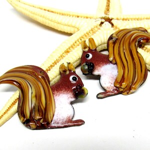 Könnte beinhalten: Zwei gläserne Eichhörnchenfiguren mit braunen, weißen und goldenen Akzenten. Jedes Eichhörnchen hält eine kleine, grüne Nuss aus Glas im Maul.